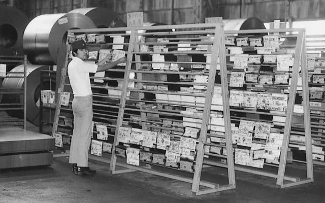 A historical black-and-white photograph of a Toyota factory worker managing inventory using an early, physical kanban board made of wooden racks and paper cards